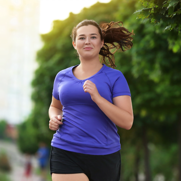 Brunette woman in a purple shirt and black shorts smiles as she jogs through a park with trees behind her.