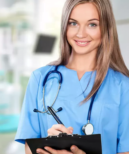 Brunette nurse looks into the camera smiling while writing on a clipboard.