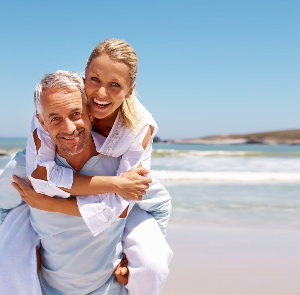 Man gives woman a piggy back ride on a beach while they are both laughing and smiling