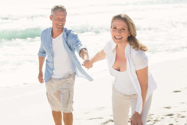 Happy couple laughs while holding hands and walking down the beach.
